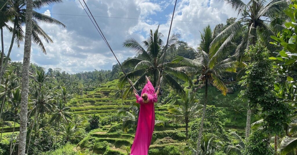 Traveler in a pink dress enjoying the famous Bali swing above Tegallalang rice terraces surrounded by lush palm trees.