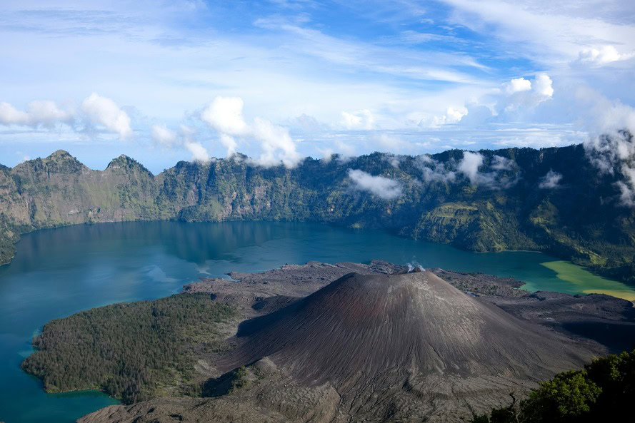 Daylight view of Mount Rinjani's volcanic cone and surrounding caldera lake on Lombok Island, Indonesia, showcasing rugged terrain and turquoise waters.